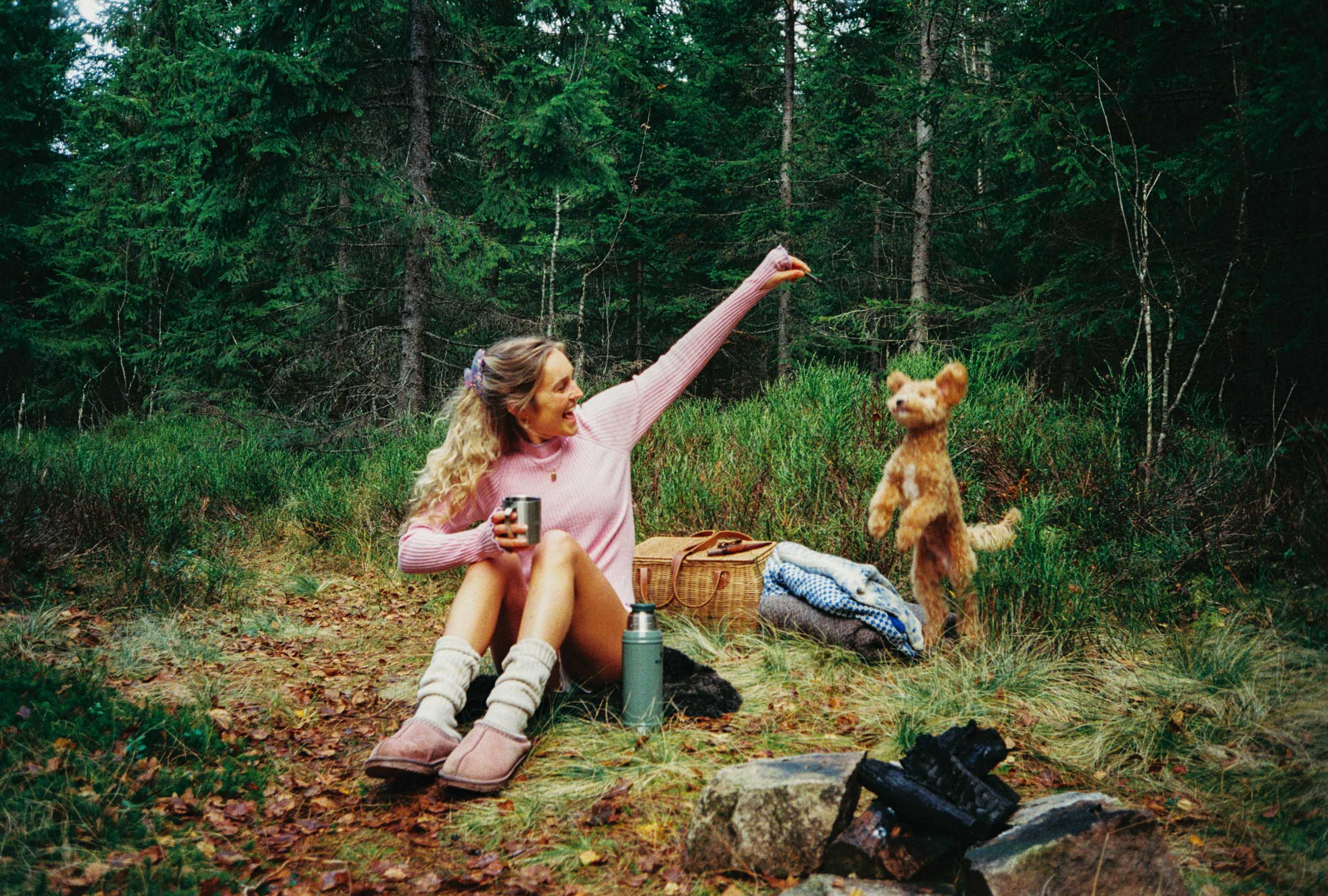 Woman sitting in a forest with a dog and picnic items, surrounded by trees.